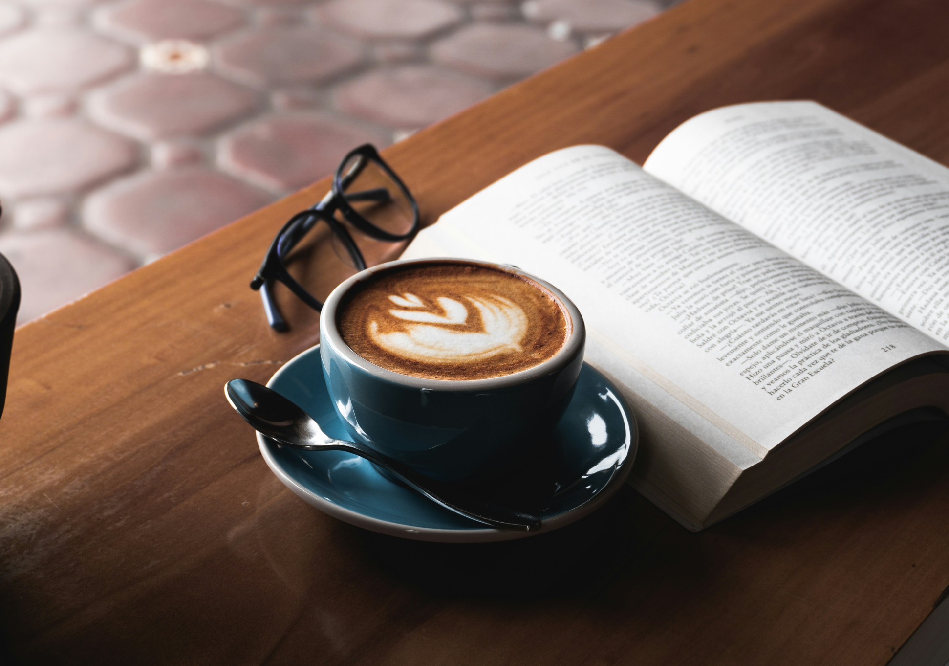 A blue cup of coffee with latte art beside an open book and reading glasses on a wooden table